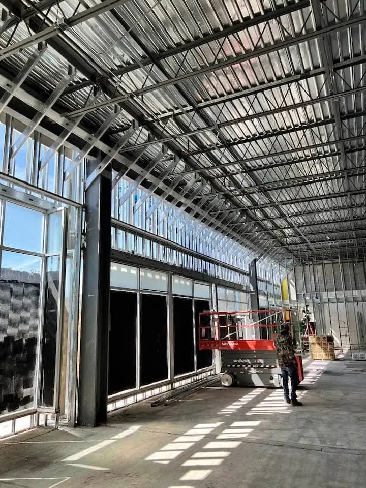 Interior of an industrial building under construction, showcasing extensive metal framework and stud work along the walls and ceiling. A worker stands near a red lift, which is positioned to facilitate access to higher areas. Large windows line one side of the building, allowing natural light to illuminate the space.