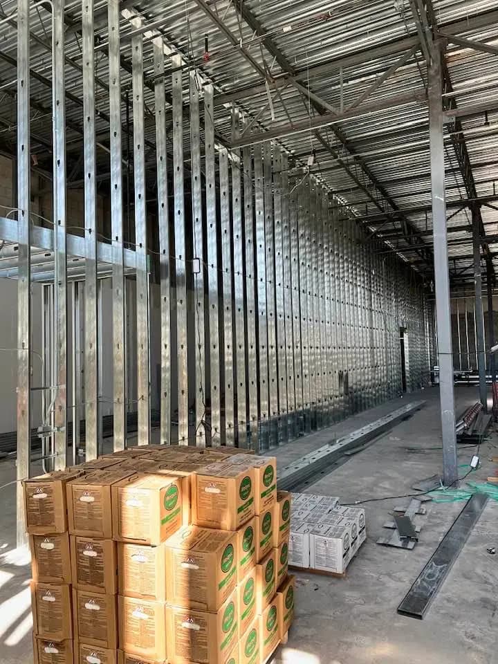 Interior of an industrial building under construction, featuring a row of vertical metal studs forming the framework for walls. Stacks of cardboard boxes are positioned in the foreground, with additional construction materials and tools scattered around the site. The exposed ceiling reveals metal beams and supports.