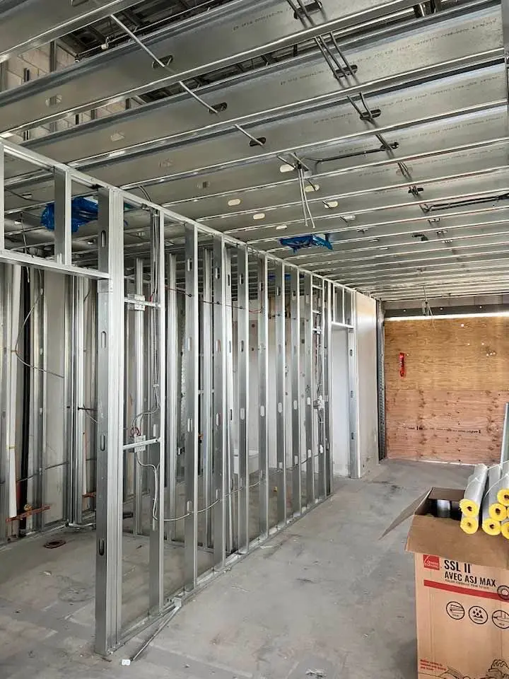 A construction site with extensive metal stud framing for walls and ceilings. The metal studs are uniformly spaced and form the skeletal structure of future rooms. Electrical wiring is threaded through some of the studs, indicating ongoing electrical work. The ceiling also features metal tracks for additional support. The floor is unfinished, emphasizing the construction phase.