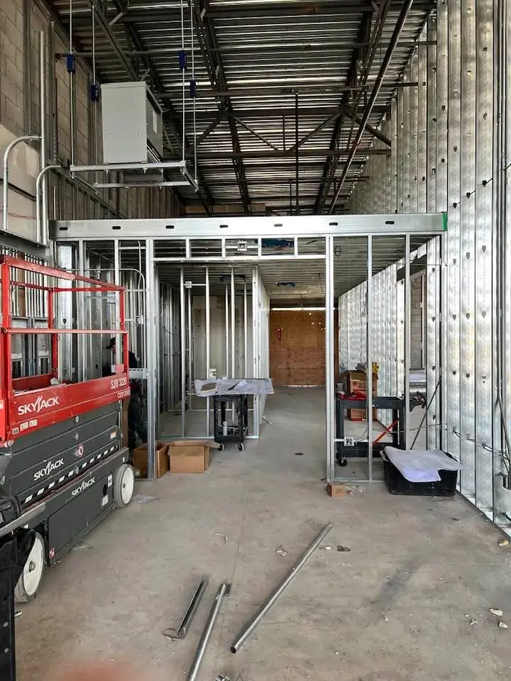 Extensive metal stud framing for both walls and ceilings. Metal studs are arranged in a grid-like pattern, forming the framework for future rooms. In the foreground, a red SkyJack lift is positioned, indicating ongoing work at height. The ceiling features exposed metal joists and an electrical box with conduit. The floor is unfinished, and various construction materials and tools are scattered throughout the area, highlighting the early stage of the build.