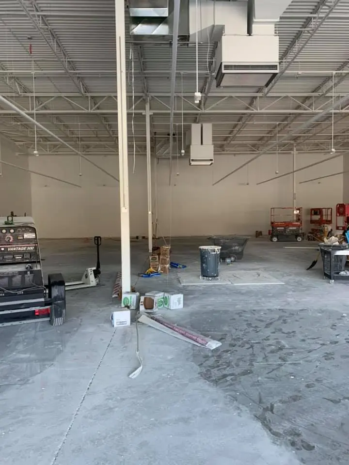 Large, empty industrial space under construction with exposed metal beams and ductwork on the ceiling, scattered construction materials and tools on the floor, and scissor lifts towards the back.