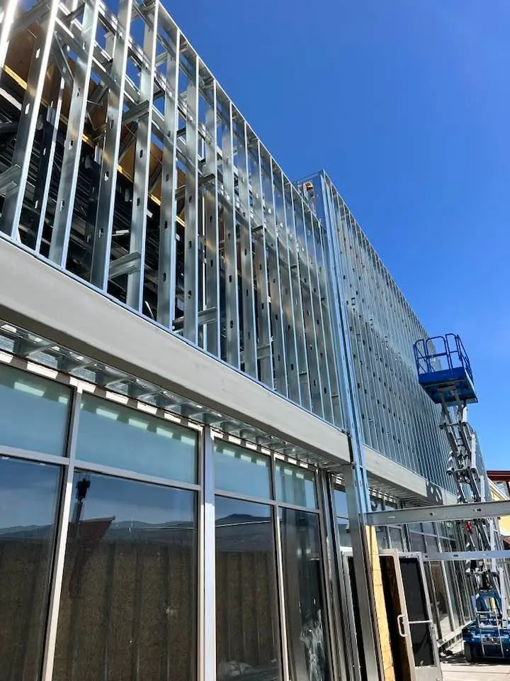 Exterior view of a building under construction with metal framing and glass windows. A scissor lift is positioned near the building, and the sky is clear and blue.