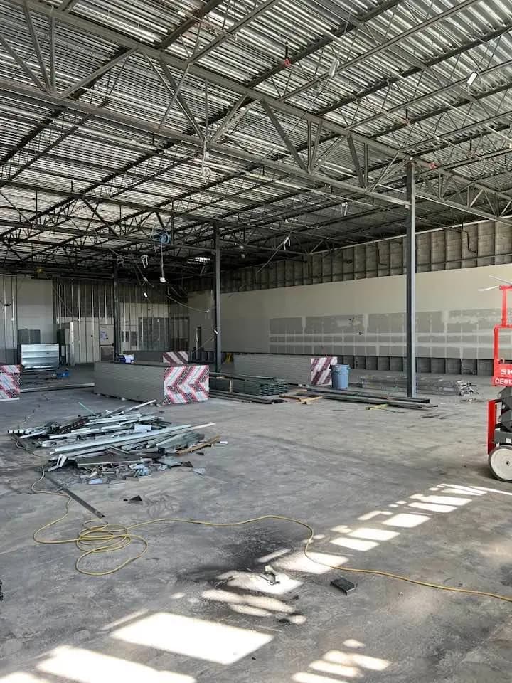 Interior view of a large industrial space under construction with exposed metal ceiling beams, scattered construction materials on the floor, and a scissor lift to the right. The walls are partially finished, and electrical wiring is visible hanging from the ceiling