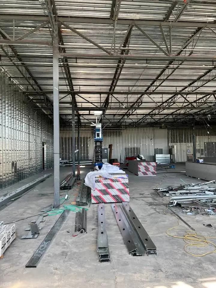 Interior of an industrial building under construction, featuring exposed metal framework, scattered construction materials, and a lift in the center. Metal stud work is visible on the floor, with steel studs and tracks laid out in preparation for creating the framework of walls and partitions.