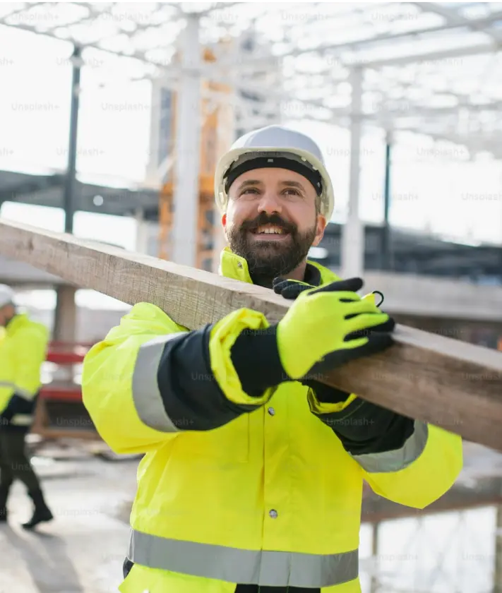 A man wearing a yellow construction outfit and helmet, holding a 4x4 piece of lumber on his left shoulder.